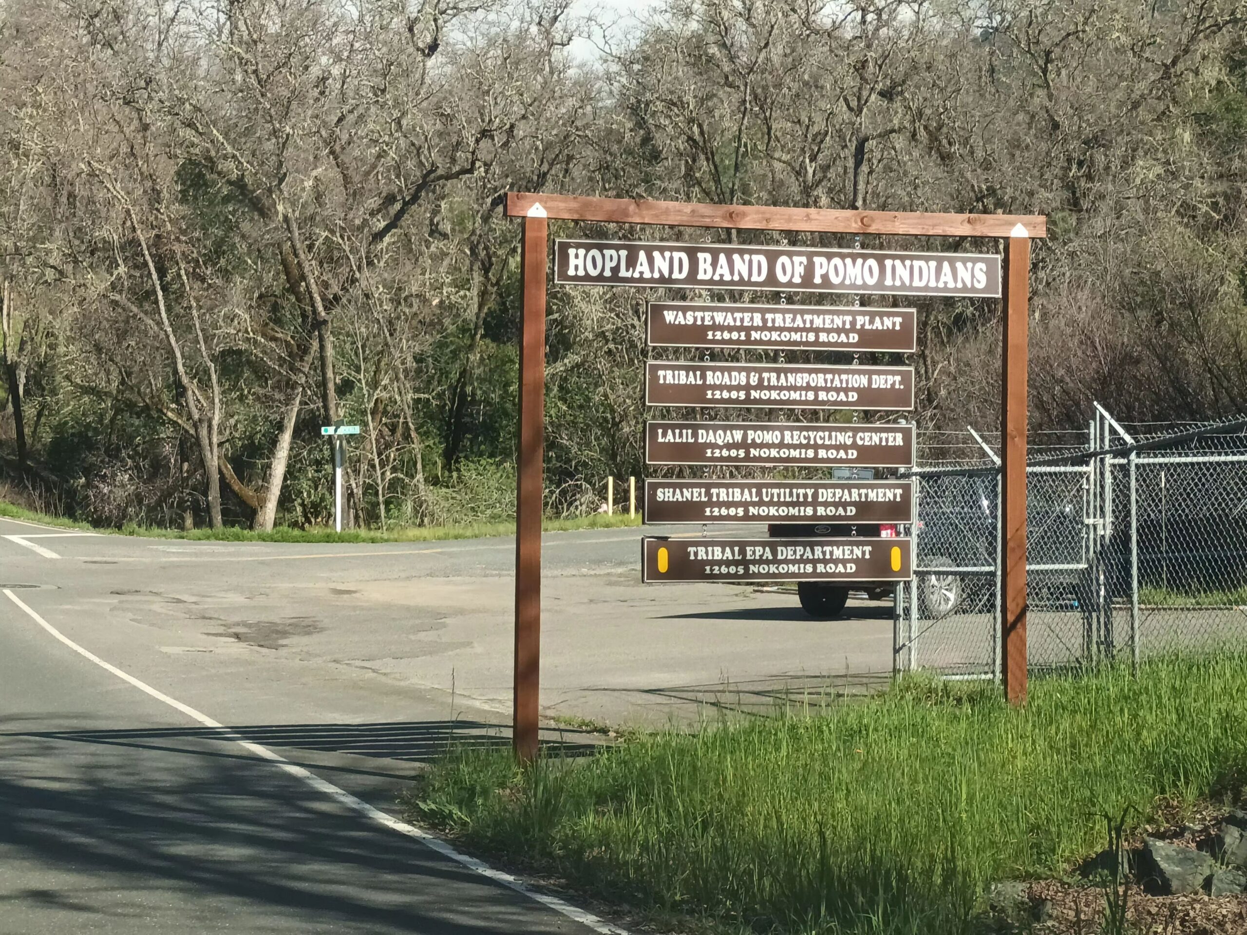 Roadside sign at the entrance to Hopland Band of Pomo Indians land, listing five departments on Nokomis Road: wastewater treatment plant, tribal roads and transportation, Lalil Daqaw Pomo recycling center, Shanel tribal utility department, and tribal EPA department.
