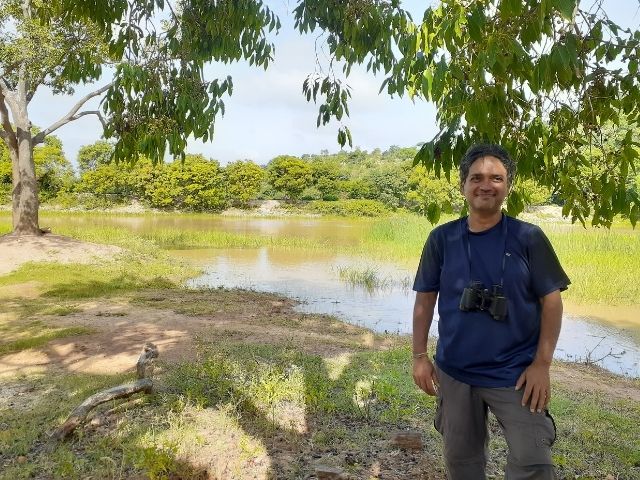 A man standing at the edge of a shallow irrigation tank in tree cover.