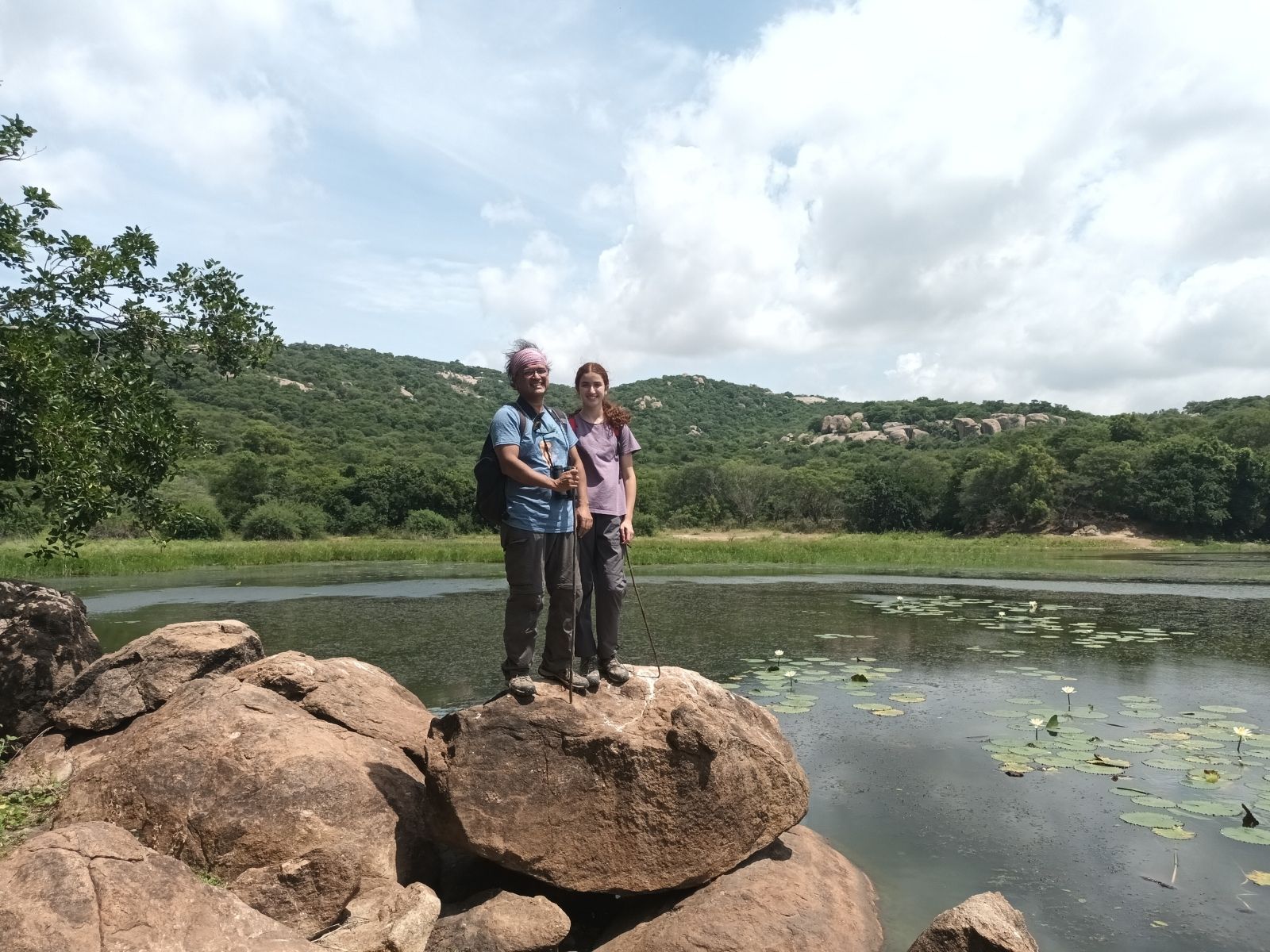 Two people standing on granite boulders at the edge of a tank covered in water lilies.