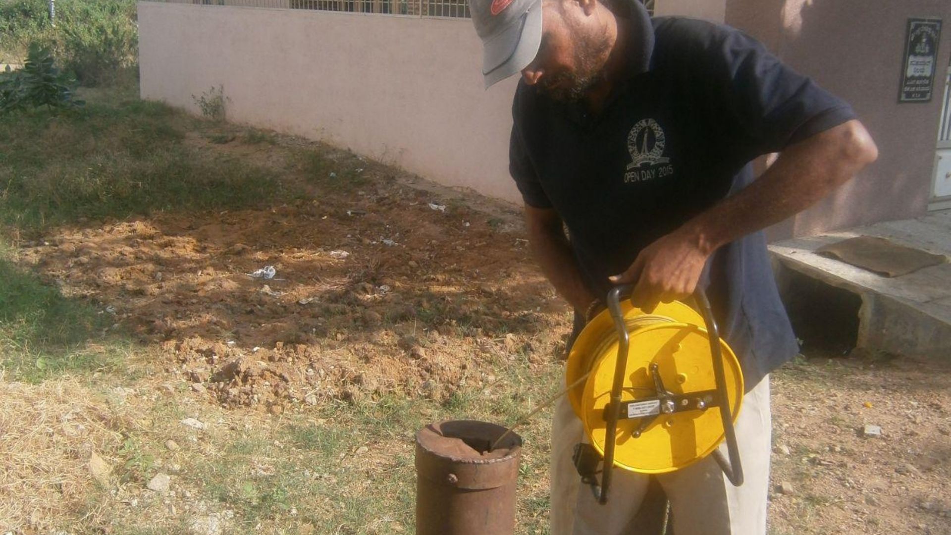 A man lowering a measuring tape into a lined borewell at the edge of a plot.